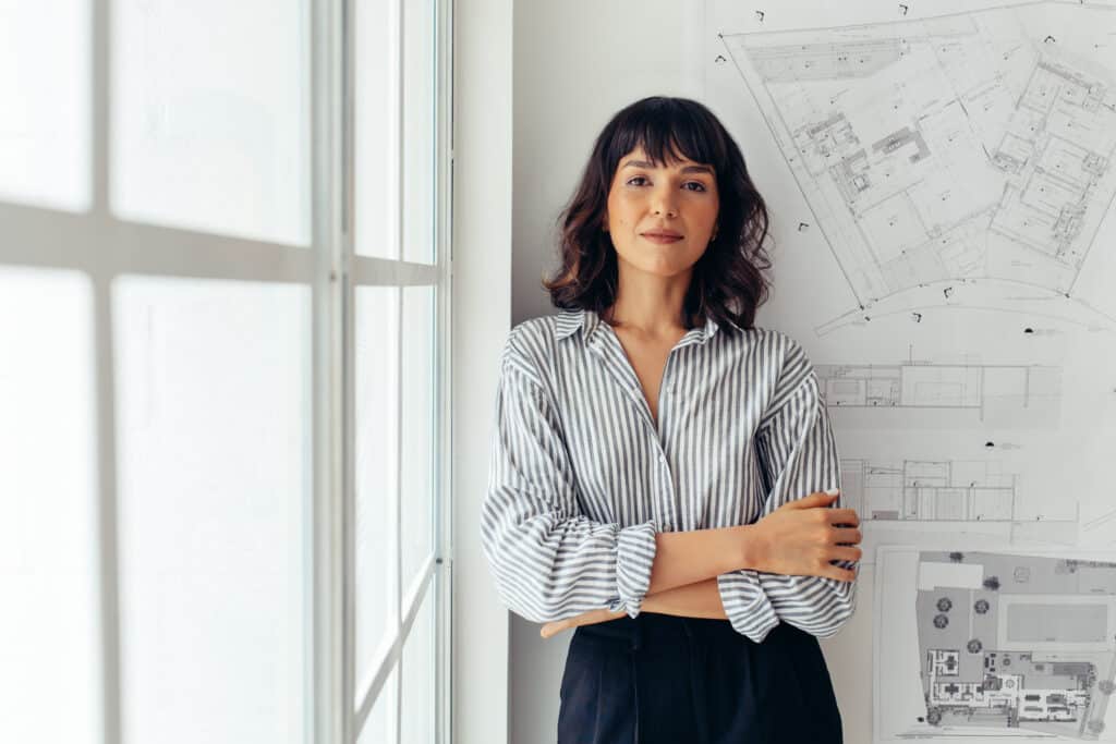 Portrait of female architect standing beside architecture drawings in office. Businesswoman standing in office with arms crossed.